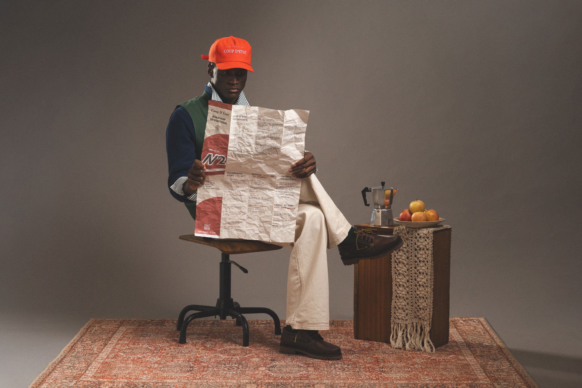 Person sitting on a chair holding a newspaper, with a small table and fruits in the background.
